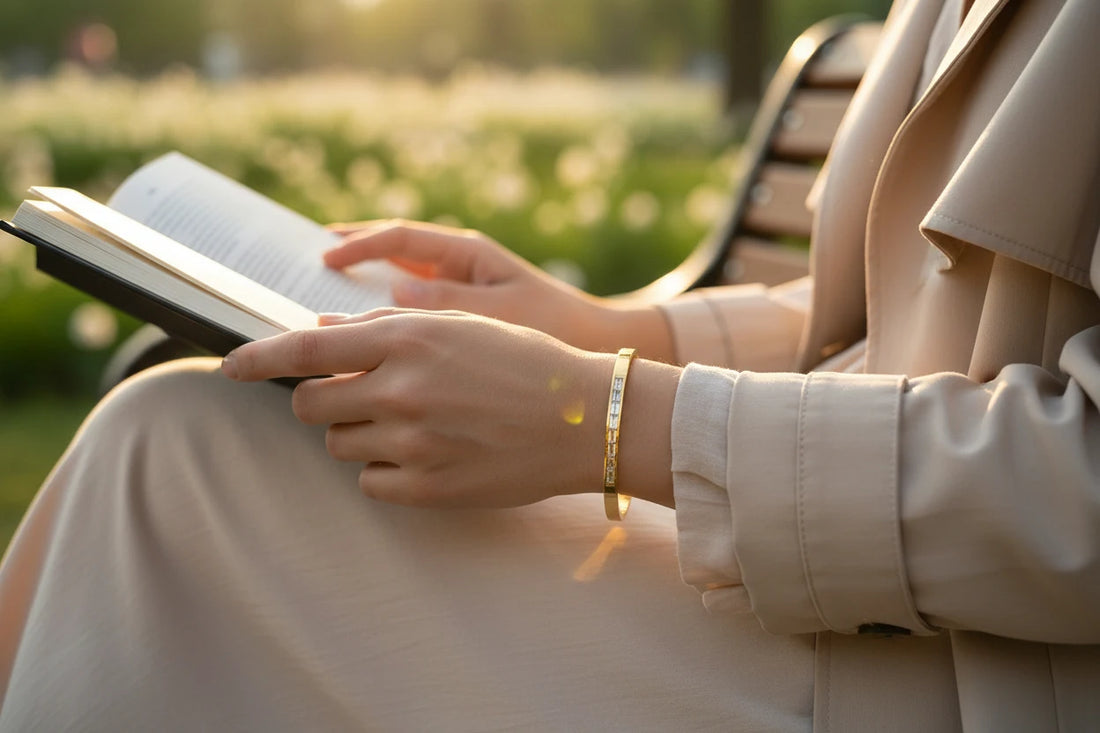 Woman reading a book on a park bench at golden hour, wearing a slim 14k gold hinged bangle bracelet that catches the sunlight against her neutral dress | Oria Jewel