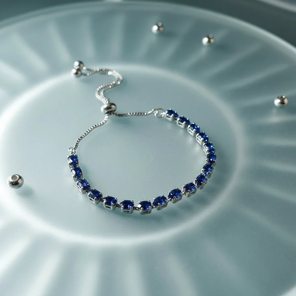 Silver Aurelia Cluster Tennis Bracelet with vivid deep-blue round crystals arranged in a neat row, styled on a frosted glass surface with soft radial light reflections and scattered metal beads for a modern, luminous look | Oria Jewel