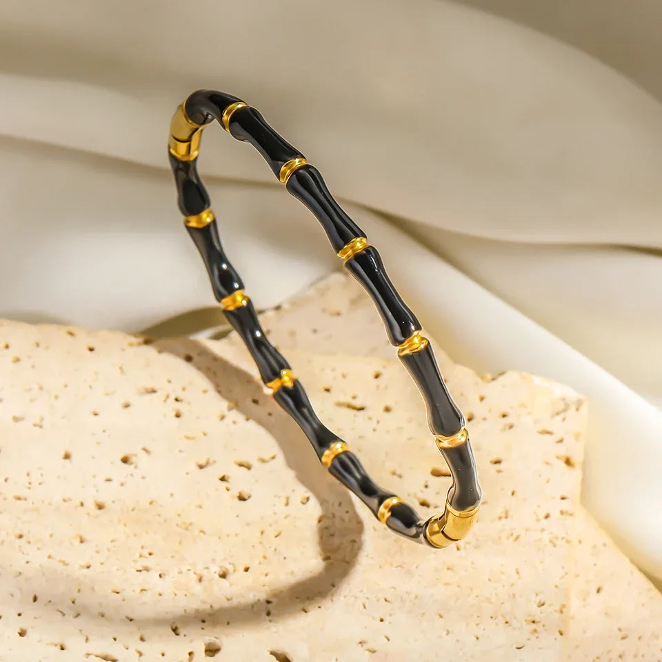 Studio shot of Black Bamboo bangle Bracelet standing upright on textured stone with draped fabric backdrop, highlighting glossy black enamel and gold rings | Oria Jewel
