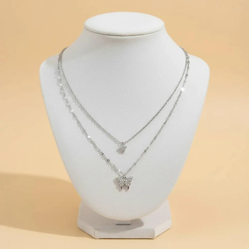 Silver Butterfly Layered Necklace displayed on a white jewelry bust against a warm beige backdrop, clearly showing both tiers and the crystal butterfly pendant | Oria Jewel