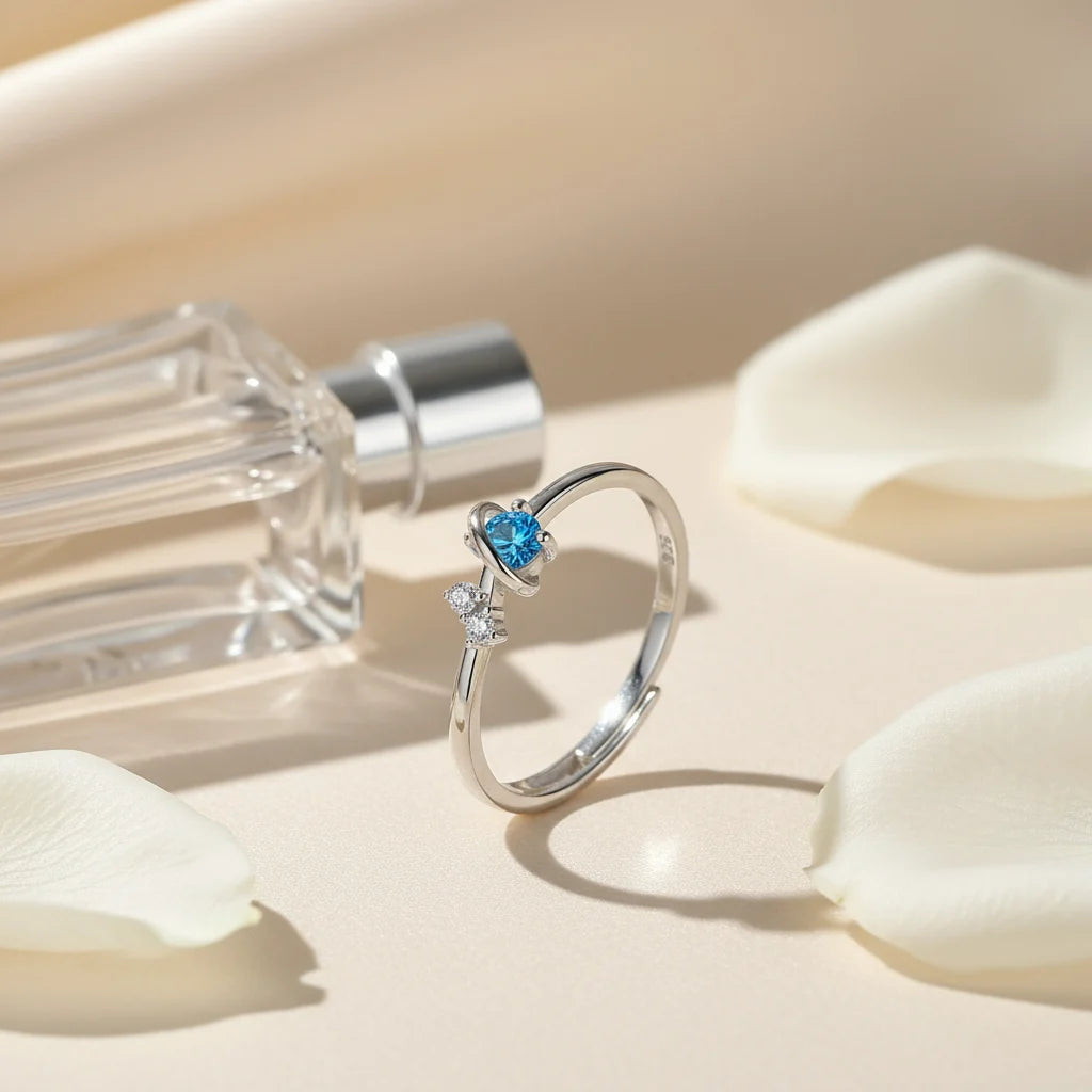 Cosmic Heart Couple Ring close-up of the open adjustable silver ring with a bright blue heart-shaped stone and tiny clear crystals, styled beside a perfume bottle and soft petals on a warm neutral surface | OriaJewel