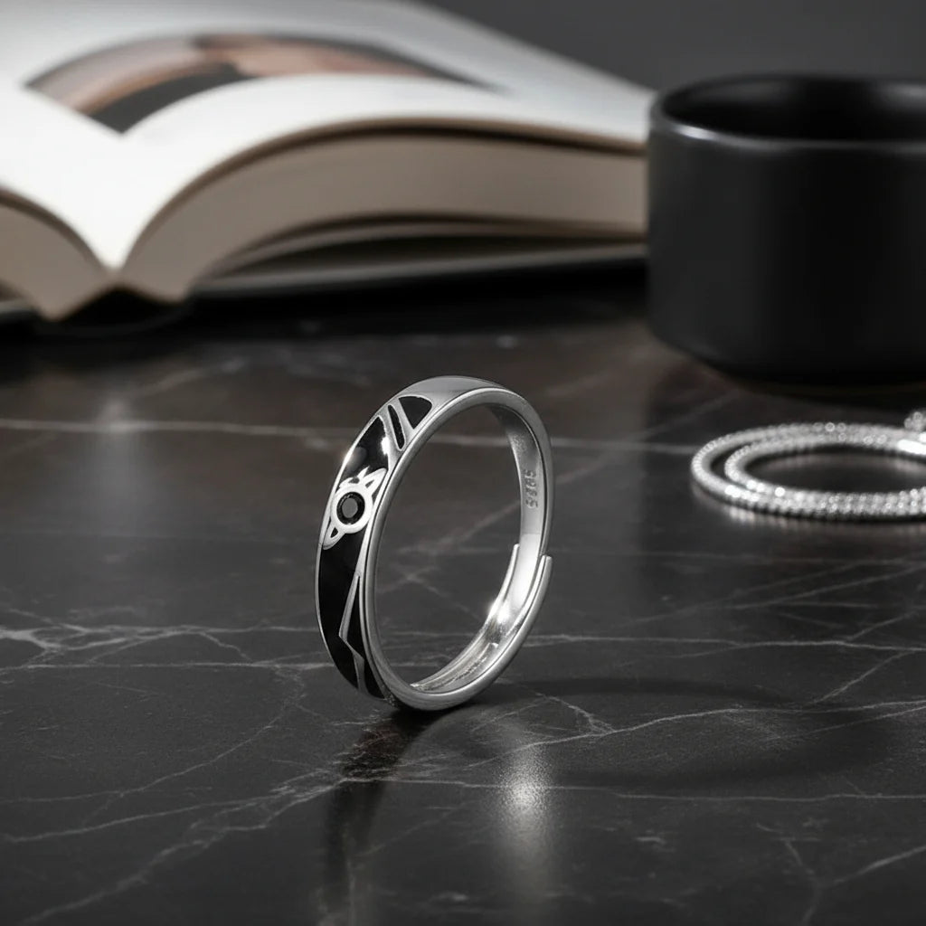 Cosmic Heart Couple Ring black enamel band ring displayed on a dark marble surface with an open book and accessories blurred behind, showcasing the planet-like motif and glossy silver finish | OriaJewel