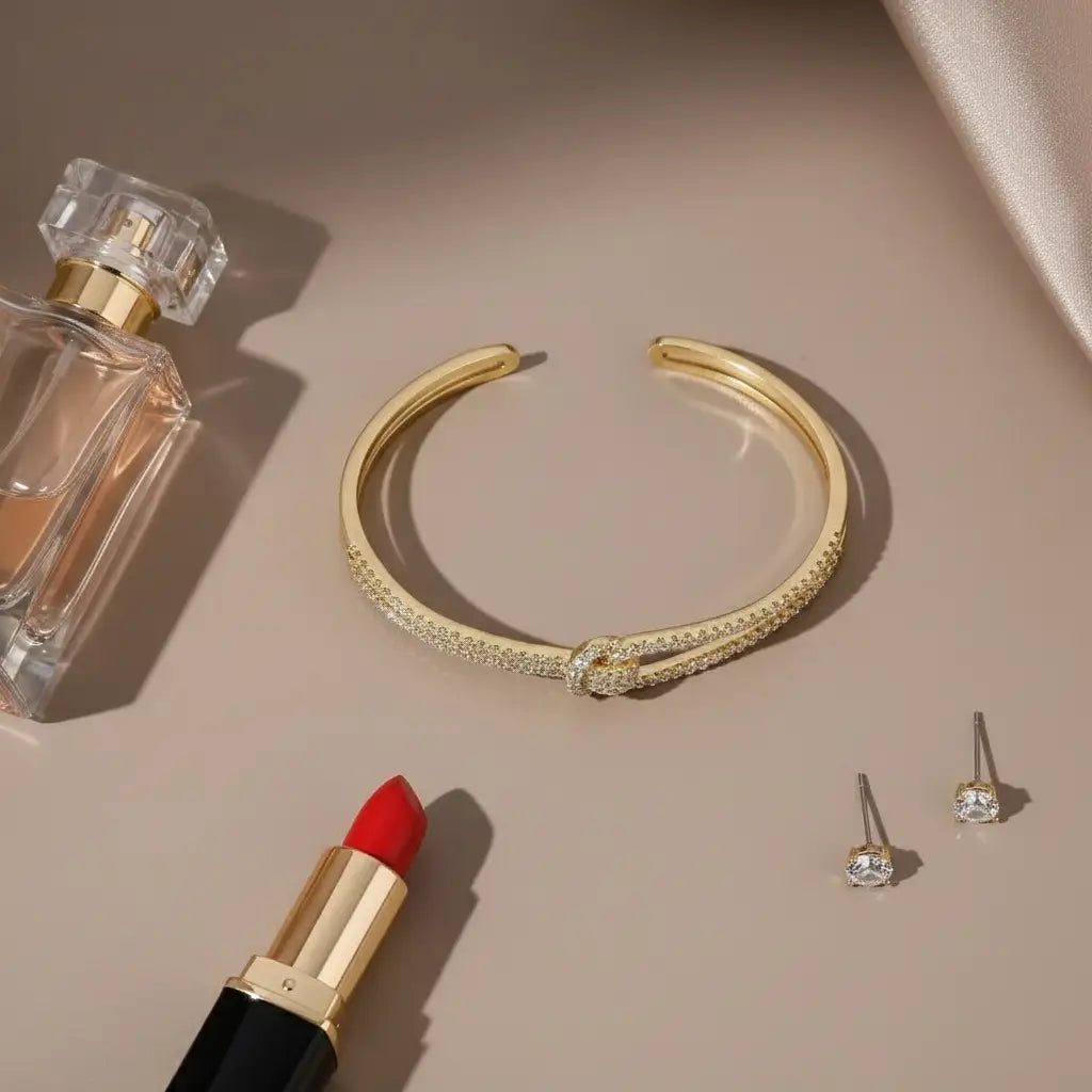 Flat lay of a gold Crystal Knot Cuff Bracelet beside perfume, red lipstick, and stud earrings on a beige background | Oria jewel