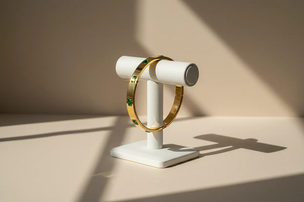 Emerald Gold Hinged Bangle displayed upright on a white T-bar stand, sunlight creating dramatic shadows and a star-like sparkle on one green stone against a soft beige backdrop | Oria Jewel