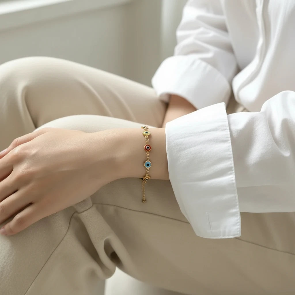 Close-up of a woman sitting with folded legs by a bright window, wearing a delicate Evil Eye Charm Bracelet with multicolored enamel charms on a gold chain, styled with a crisp white shirt and beige trousers | Oria Jewel