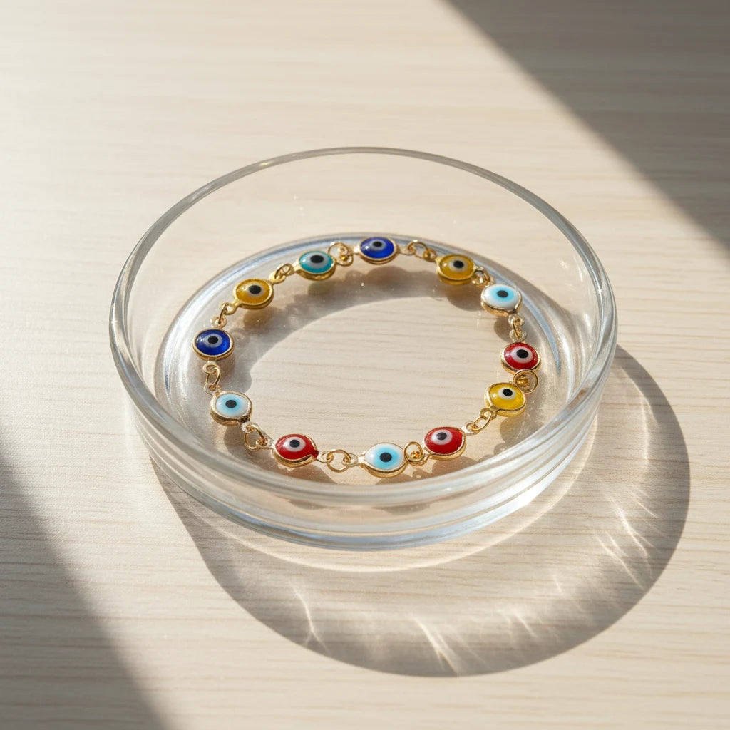 Sunlit close-up of the Evil Eye Charm Bracelet arranged inside a clear glass dish on a wooden surface, the colorful glass eye charms glowing softly in the natural light for a bright, uplifting vibe | Oria Jewel