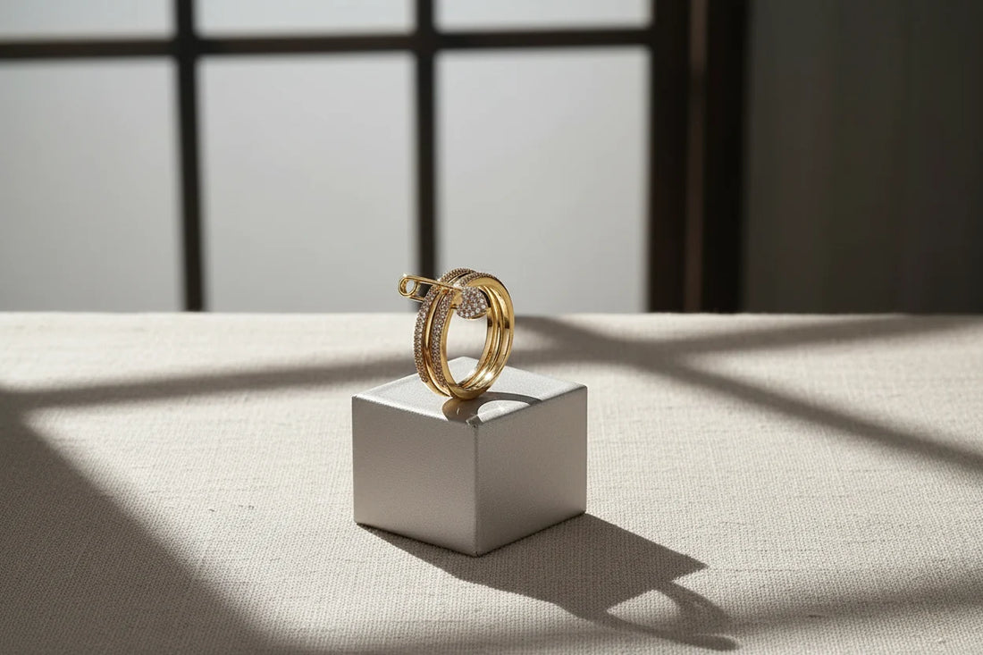 Gold Safety Pin Ring displayed on a small cube pedestal with soft window shadows, minimal editorial scene, gold-tone pavé bands with safety pin detail and pavé heart charm | OriaJewel