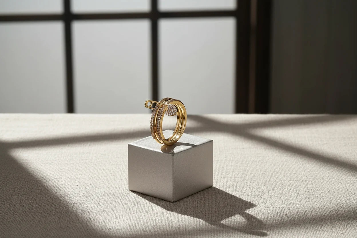 Gold Safety Pin Ring displayed on a small cube pedestal with soft window shadows, minimal editorial scene, gold-tone pavé bands with safety pin detail and pavé heart charm | OriaJewel
