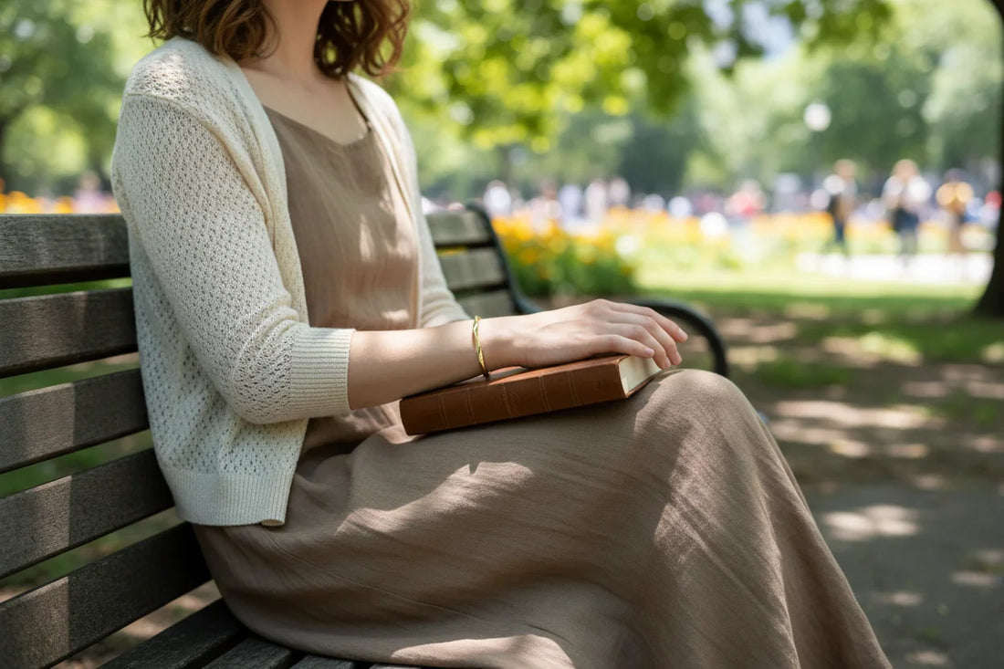 Woman relaxing on a park bench under soft daylight, wearing a Gold twisted cuff bracelet that complements her light dress and knitted cardigan while resting her hand on a closed book | Oria Jewel