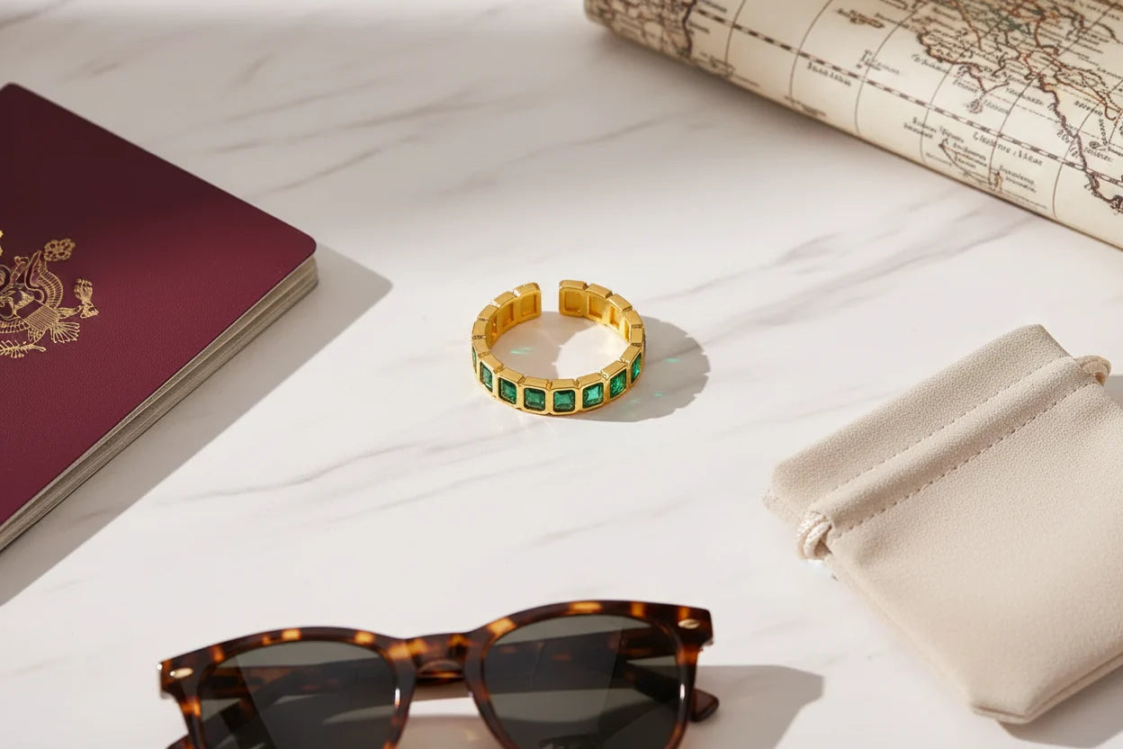 Green stone band ring flat lay on a bright marble surface with travel accessories, sunglasses and a pouch nearby, sharp detail on the gold-tone band and green stones | OriaJewel