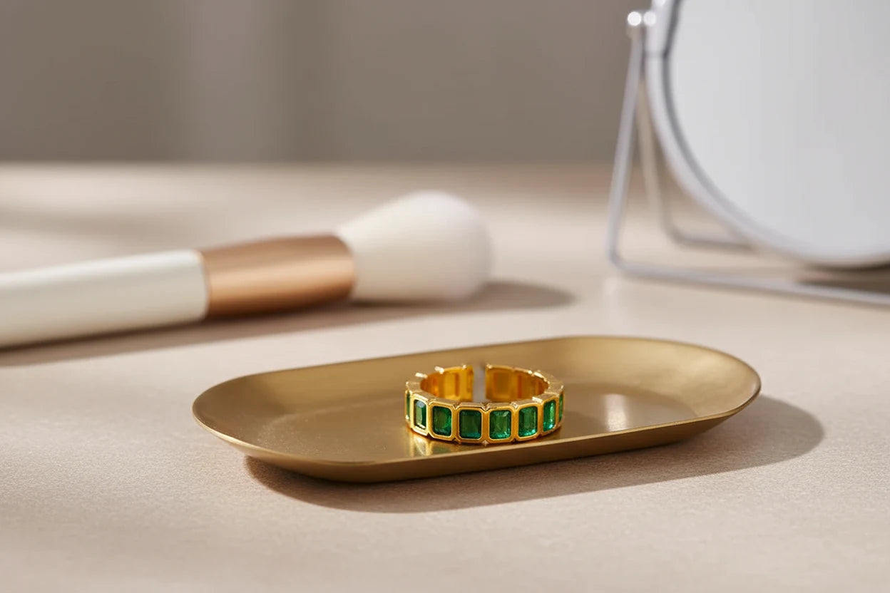 Green stone band ring placed on a brushed gold tray near beauty tools and a mirror, warm editorial lighting and close focus on the gold-tone finish and green stones | OriaJewel