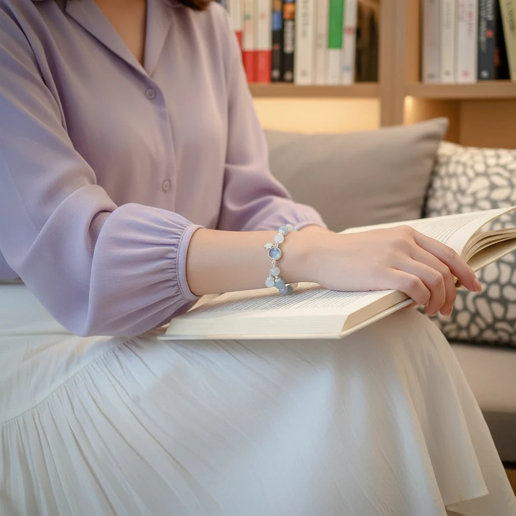 Woman reading on a sofa wearing a pastel blouse and white skirt, showing the delicate Moonstone Glow Charm Bracelet on her wrist | Oria Jewel