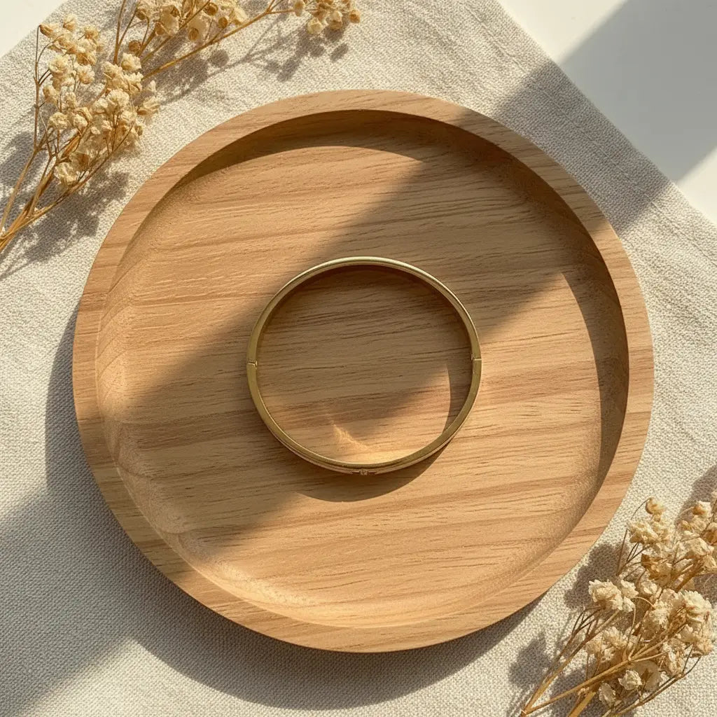 Top view of the Mother of Pearl Gold Bangle resting inside a round wooden tray, warm sunlight and dried flowers creating a cozy, natural scene that emphasizes the braceletโs golden shine | Oria Jewel