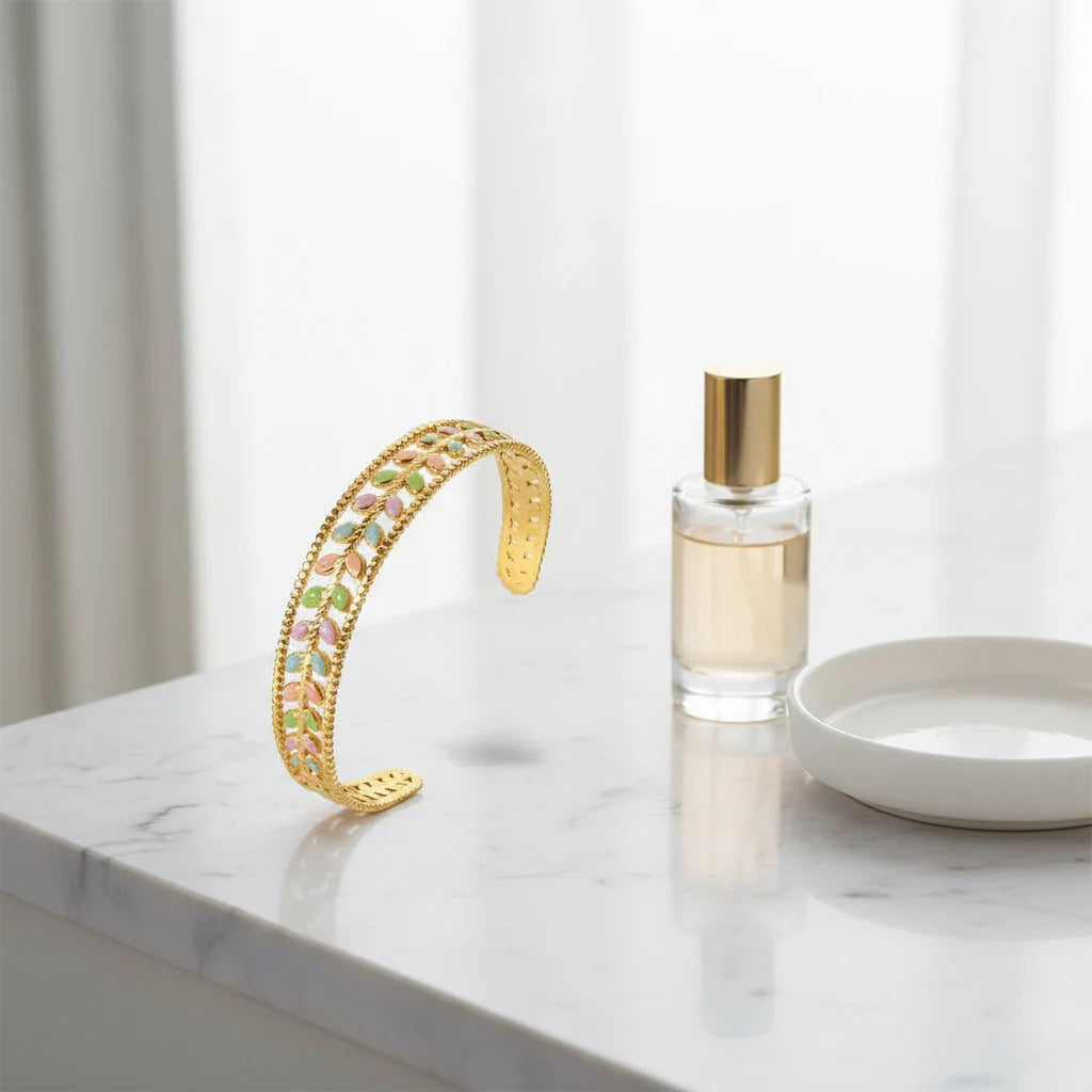 Airy bathroom counter shot of the Pastel Leaf Garden Cuff Bracelet standing upright near a perfume bottle and dish on white marble, focusing on the open cuff silhouette and delicate pastel leaf motif | Oria Jewel