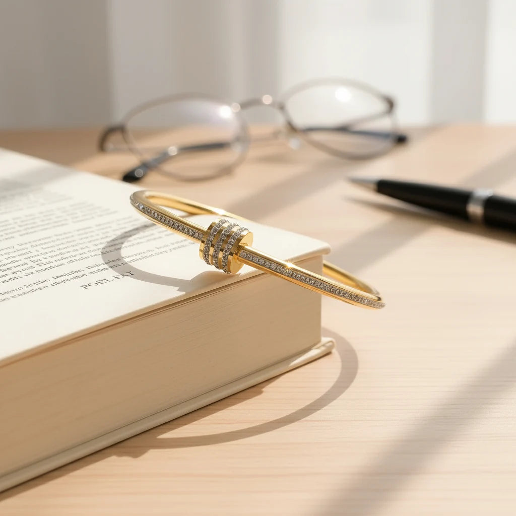 The Pavé Stackable Gold Bangle balanced on the edge of an open book on a wooden desk, with reading glasses and a pen in the background, suggesting a sophisticated everyday piece that transitions from work to leisure | Oria Jewel