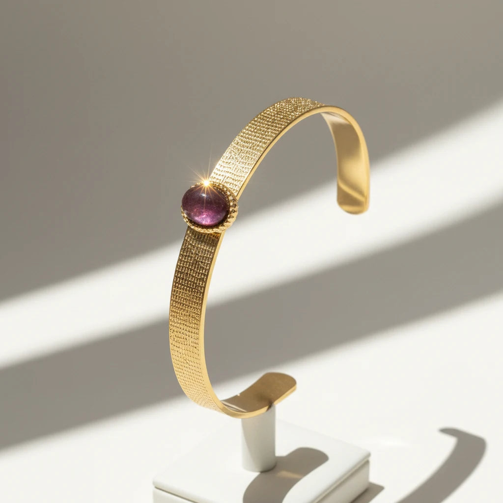 Plum gemstone cuff bracelet displayed upright on a small white stand, with sunlight creating bright highlights on the textured gold band and sparkling off the domed plum stone. | Oria Jewel
