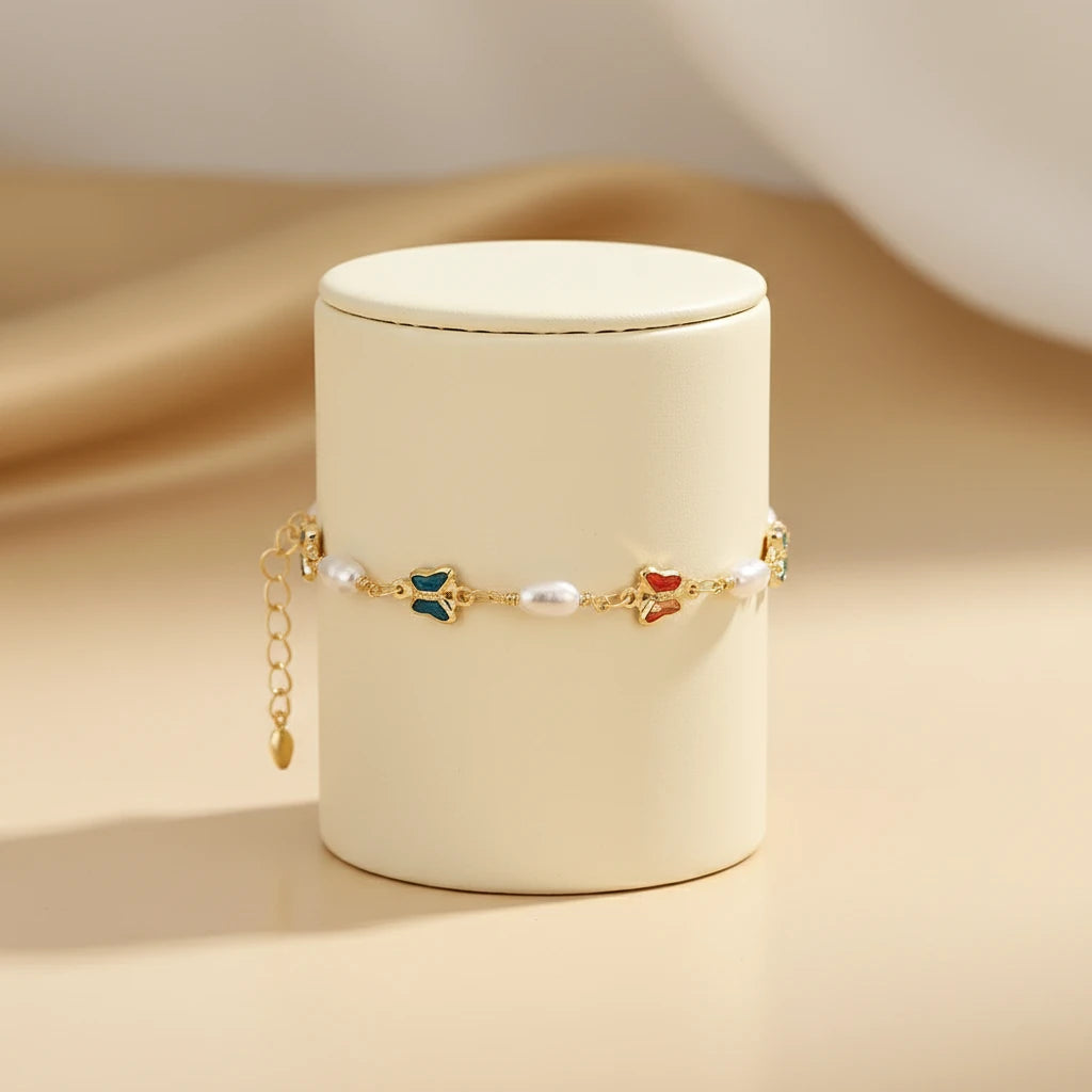 Rainbow Butterfly Pearl Chain Bracelet displayed wrapped around an elegant ivory cylinder stand, showing its gold chain with colorful enamel butterfly charms and small white pearls against a soft beige backdrop | Oria Jewel