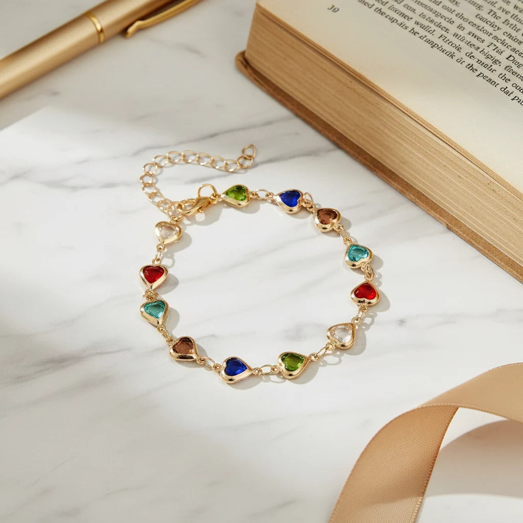 Rainbow Heart Charm Bracelet laid flat on a white marble surface next to a book and gold pen, showcasing each tiny multicolor heart link in detail | Oria Jewel