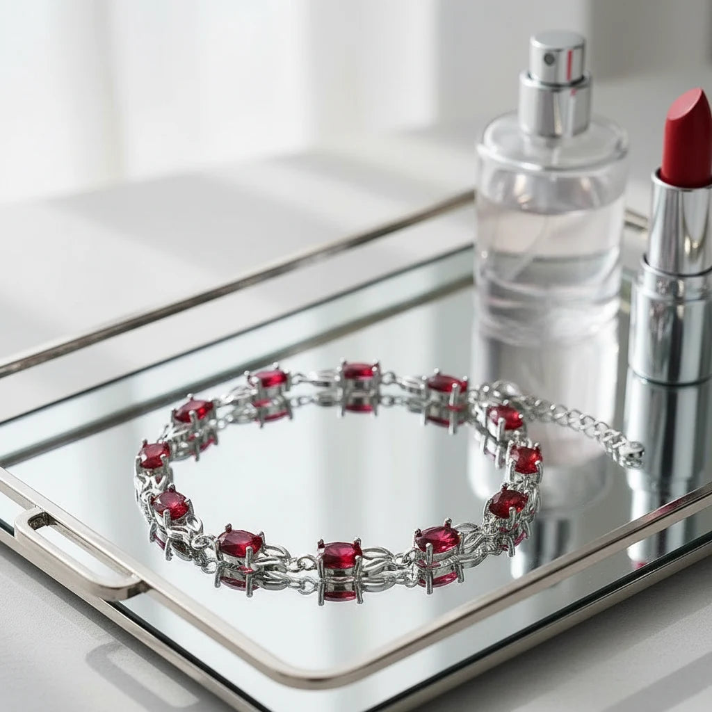 Red Crystal Silver Tennis Bracelet placed on a mirrored tray beside a clear perfume bottle and red lipstick, with reflections highlighting the vivid red stones and intricate silver chain design | Oria Jewel