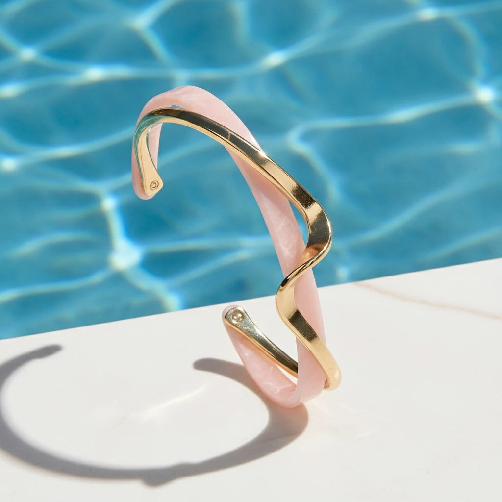 Rosette twist cuff bracelet in pink enamel and gold balanced on a white poolside ledge, with sparkling blue water reflections emphasizing its wavy summer-ready design | Oria Jewel