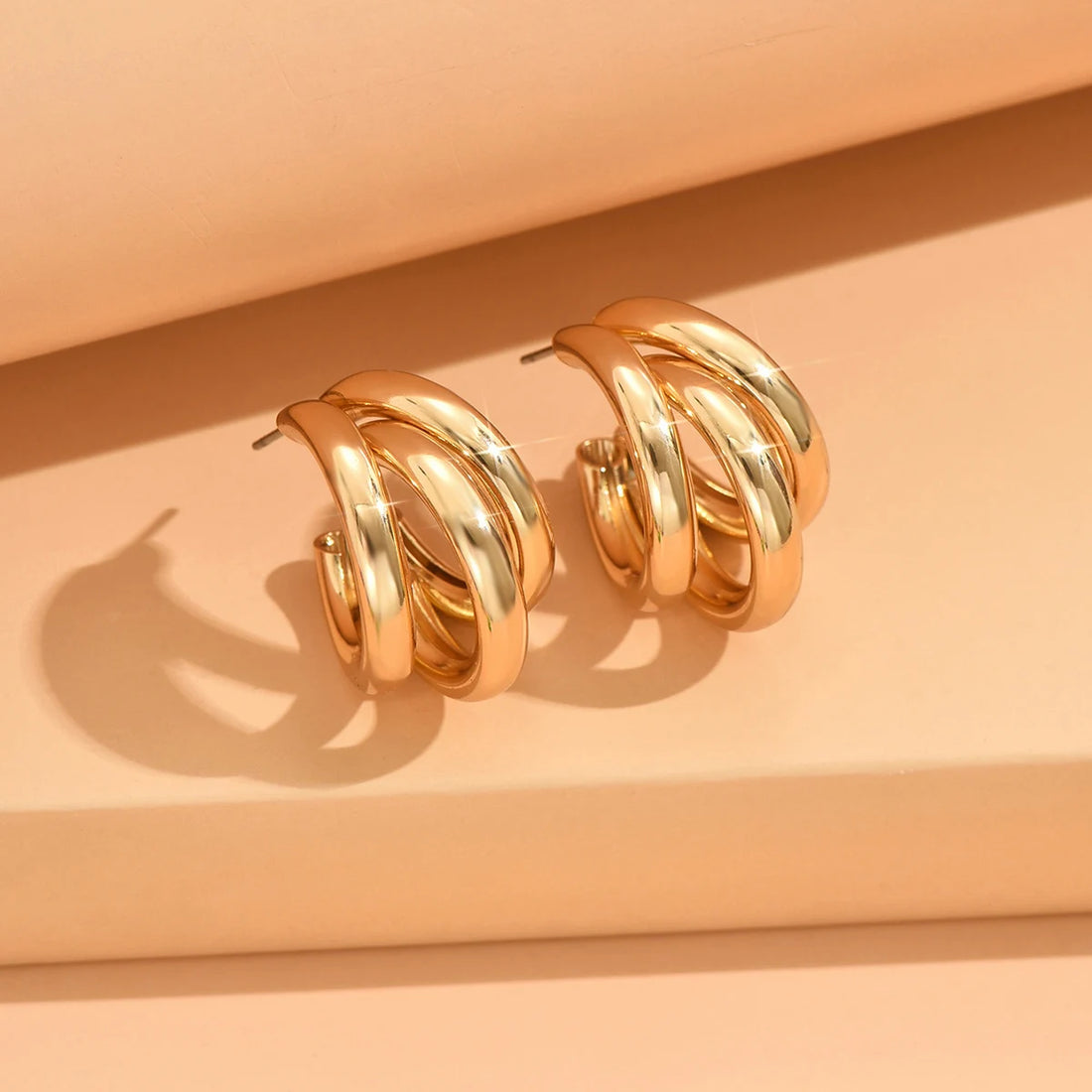 Shiny gold geometric hoop earrings on a peach surface, casting shadows | Oria Jewel