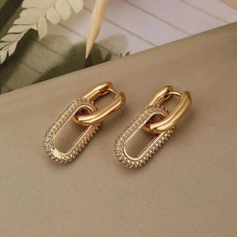 Gleaming gold little hoops with textured oval links on a beige surface. | Oria Jewel