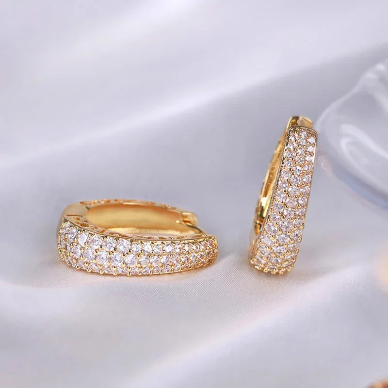 Two brilliant Crystal Gold Huggie Earrings gleam on a soft, folded white fabric | Oria Jewel