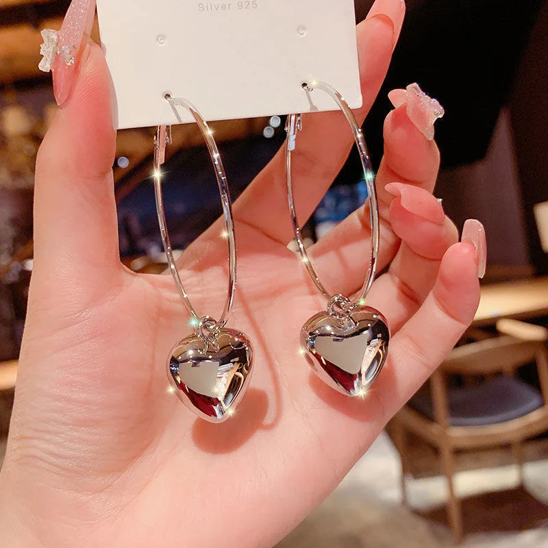 A hand holds shimmering silver heart drop earrings, reflecting light brightly | Oria Jewel