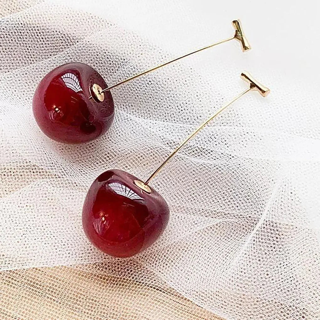 Shiny red cherry drop earrings with gold posts resting on sheer white fabric | Oria Jewel