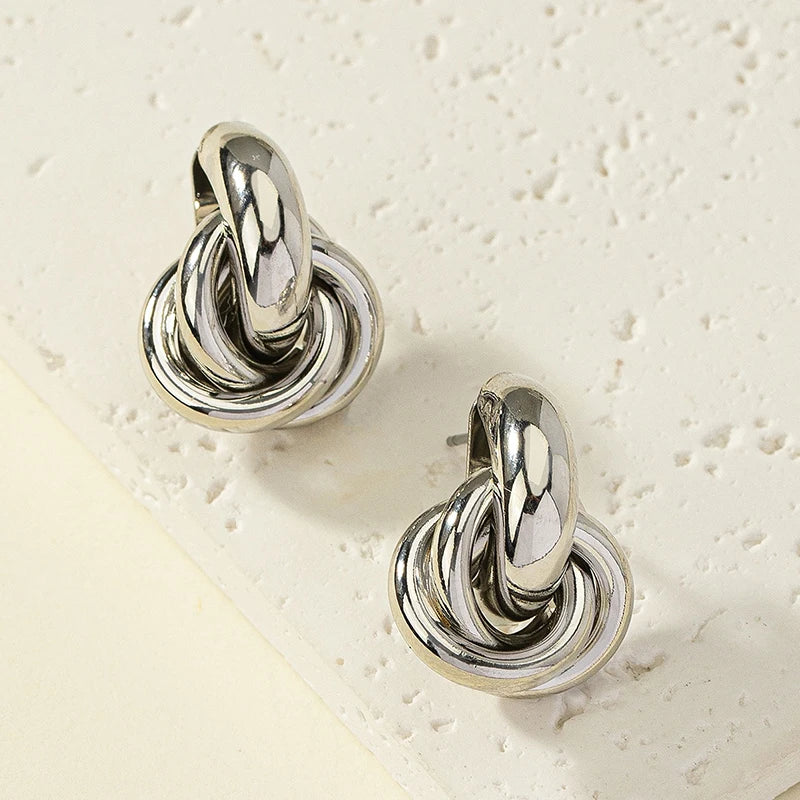 Two shiny, silver-toned Elegant Knot Hoop Earrings on a textured cream surface | Oria Jewel