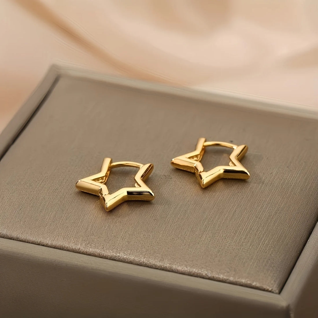 Two polished gold small hoop star earrings gleam on a textured taupe surface | Oria Jewel