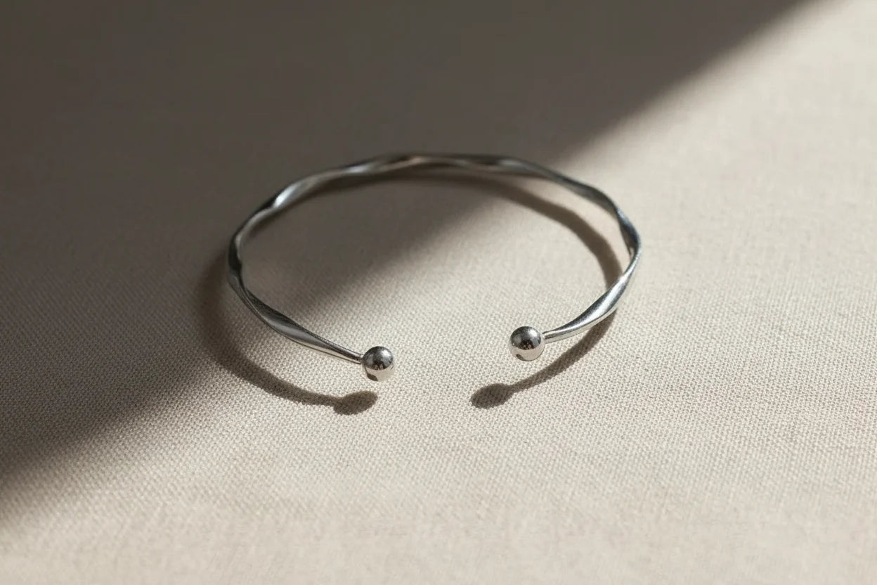 Silver twisted cuff bracelet lying open on a taupe fabric surface, sunlight casting gentle shadows that accentuate its twisted design and round ends | Oria Jewel