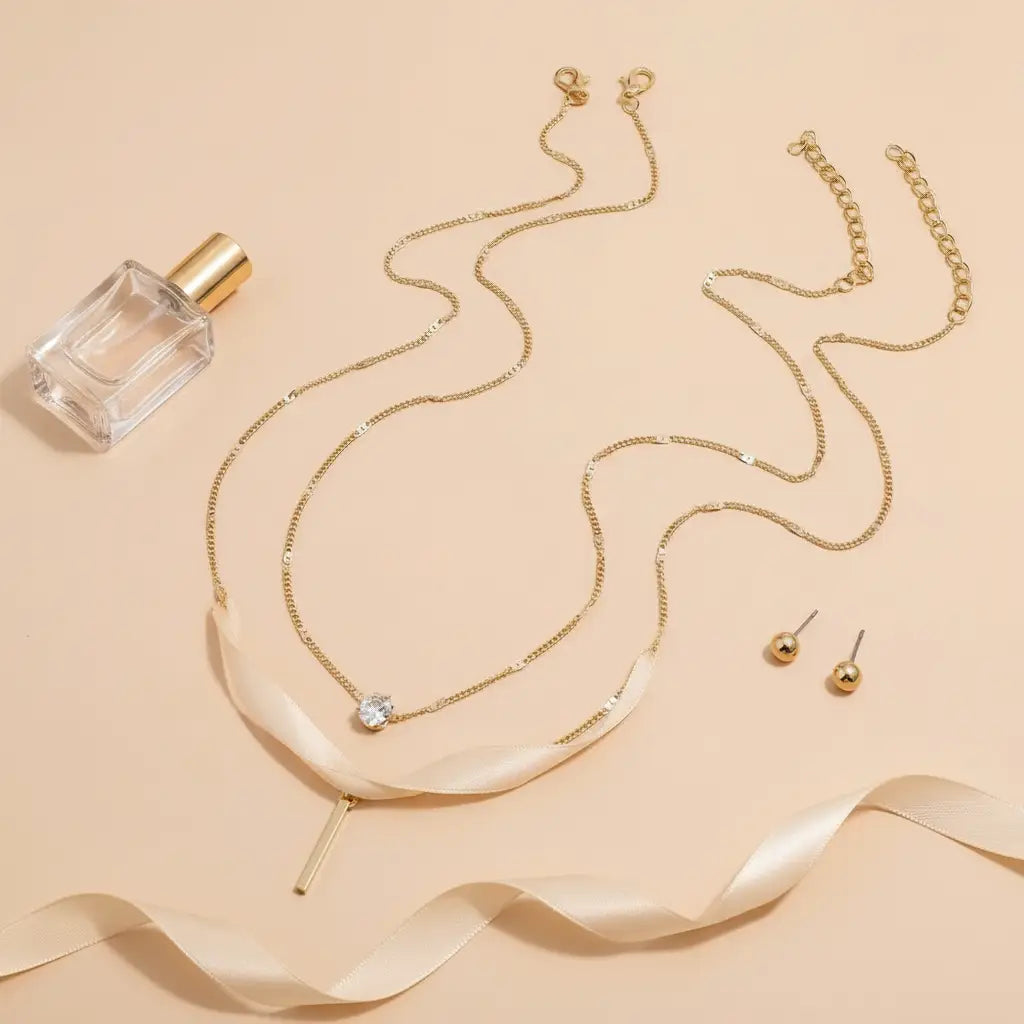 Flat lay of gold Star Layered Necklace arranged with matching gold stud earrings, ribbon, and perfume bottle on peach background | Oria Jewel