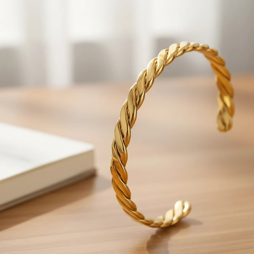 Detailed close-up of a Twisted Gold Cuff Bracelet standing upright on a wooden desk next to an open book, the smooth twisted strands and reflective gold finish highlighted by soft daylight | Oria Jewel