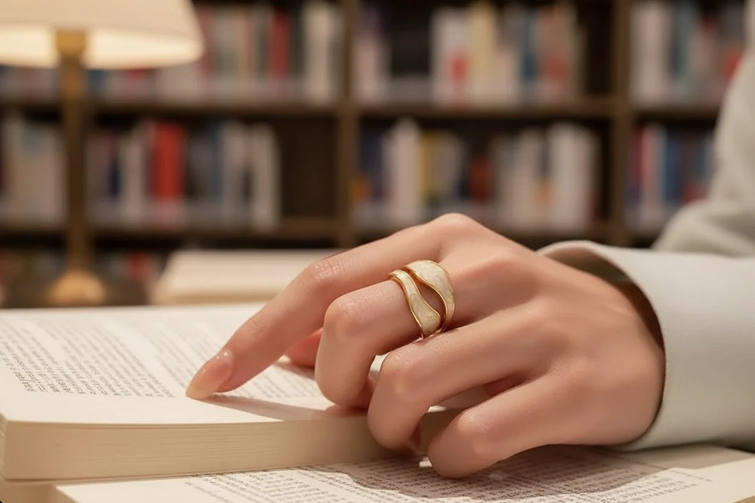 White Enamel Open Wave Ring worn while turning pages of a book in a cozy library setting, showcasing the gold-tone wavy bands layered over glossy white enamel | OriaJewel