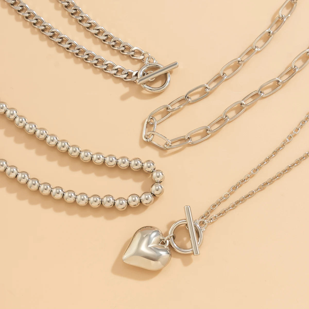Y2K Heart Layered Necklace in silver with chunky chain, bead, and link layers arranged on beige background | Oria Jewel