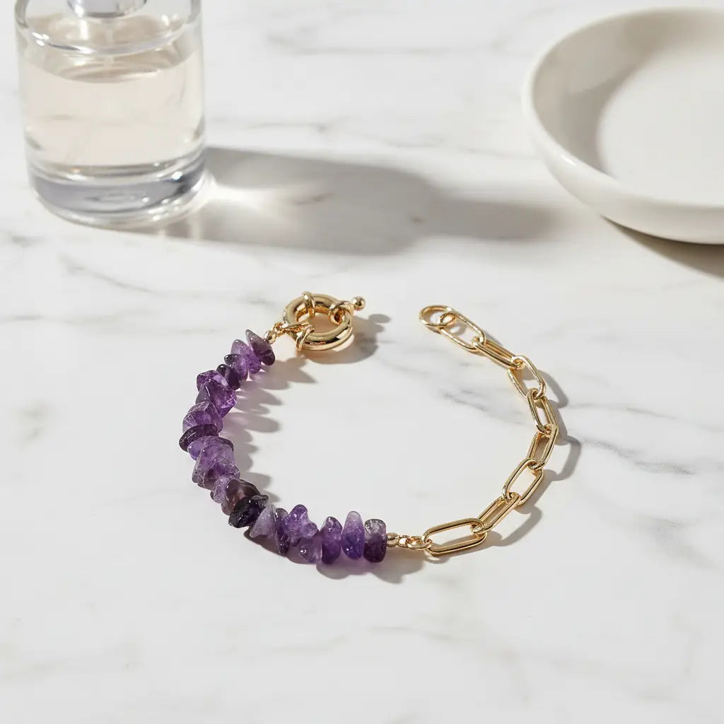 Close-up of a gold paperclip chain and purple gemstone chip amethyst bead bracelet resting on a white marble surface beside a clear perfume bottle and ceramic dish, softly lit to highlight the textured stones and polished metal | Oria Jewel