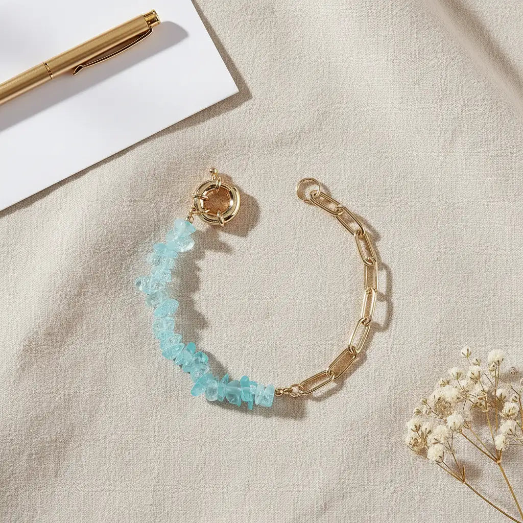 Gold paperclip chain bracelet with icy light blue gemstone chips in the same modern amethyst bead bracelet design, styled on a neutral beige fabric background with a gold pen and delicate dried flowers for an airy, elegant look | Oria Jewel
