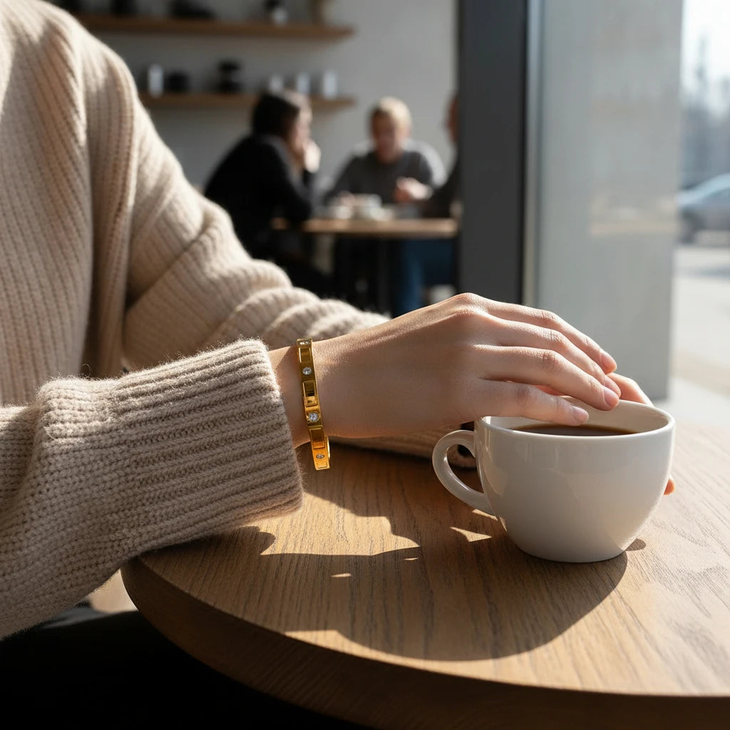 Cozy lifestyle shot of a woman in a beige sweater holding a coffee cup at a wooden café table, wearing the gold crystal cuff bracelet on her wrist | Oria Jewel