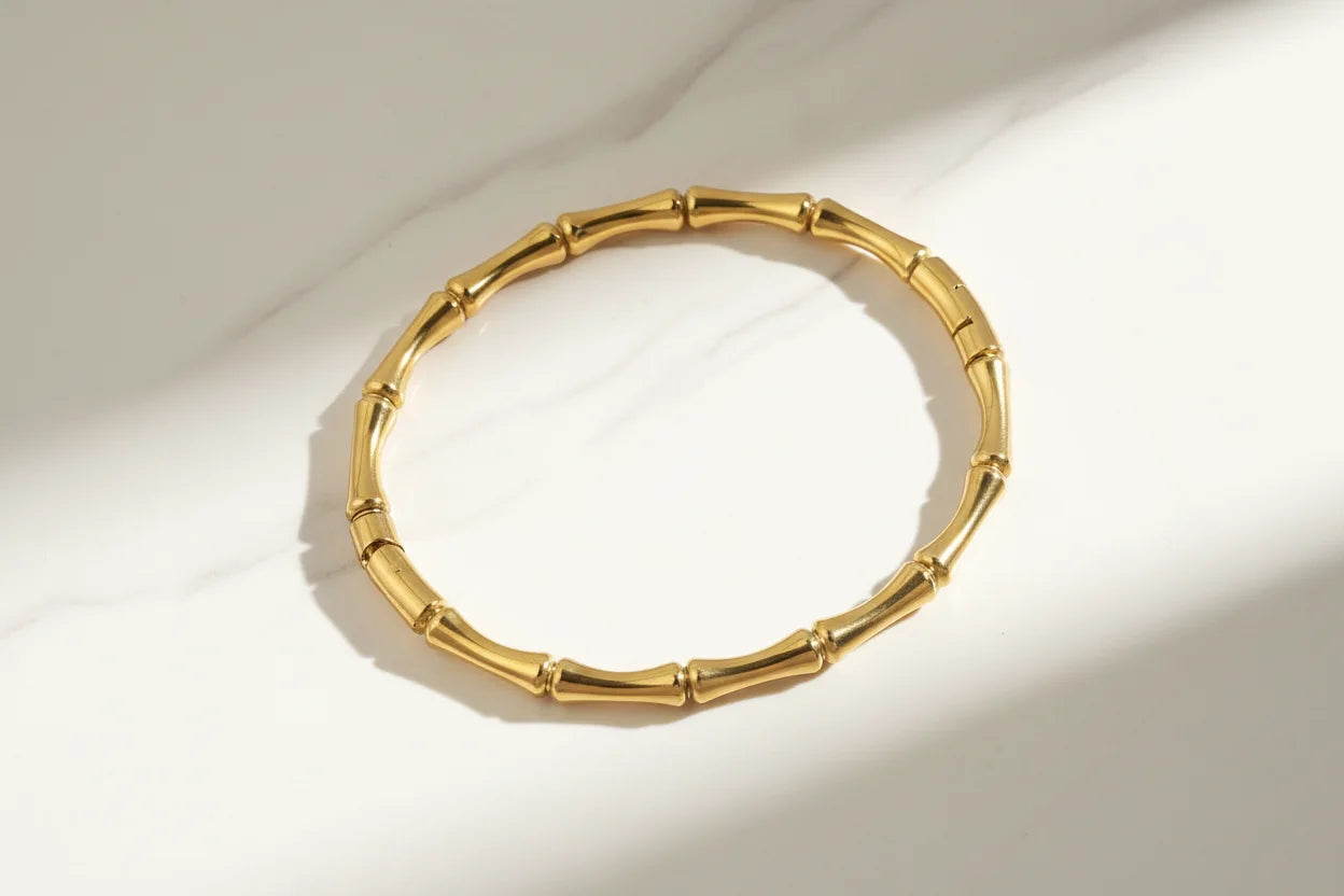 Solo product shot of a shiny gold Bamboo bangle Bracelet laid flat on white marble, highlighted by natural sunlight and soft shadows to show its curved bamboo links | Oria Jewel