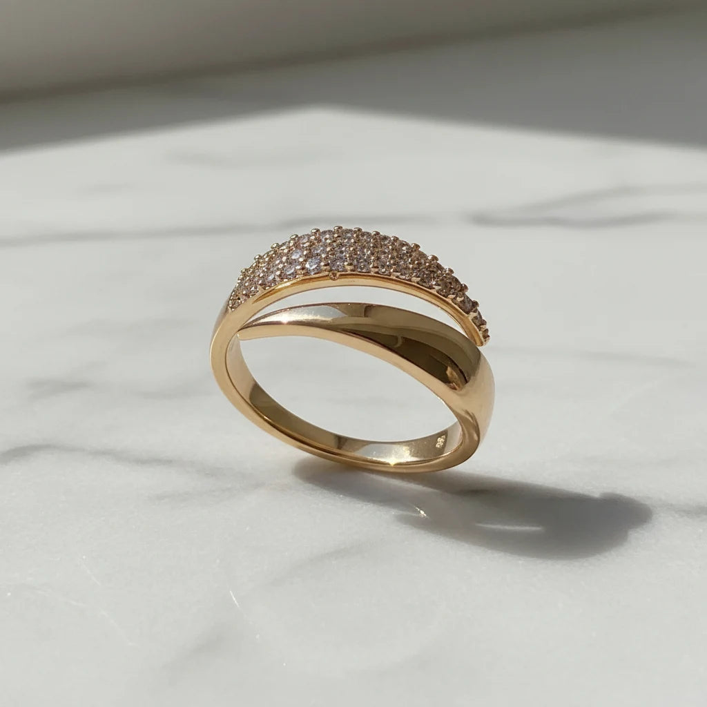 Product shot on white marble with natural shadow, featuring a glossy curved gold bypass ring and its pavé sparkle in daylight | OriaJewel