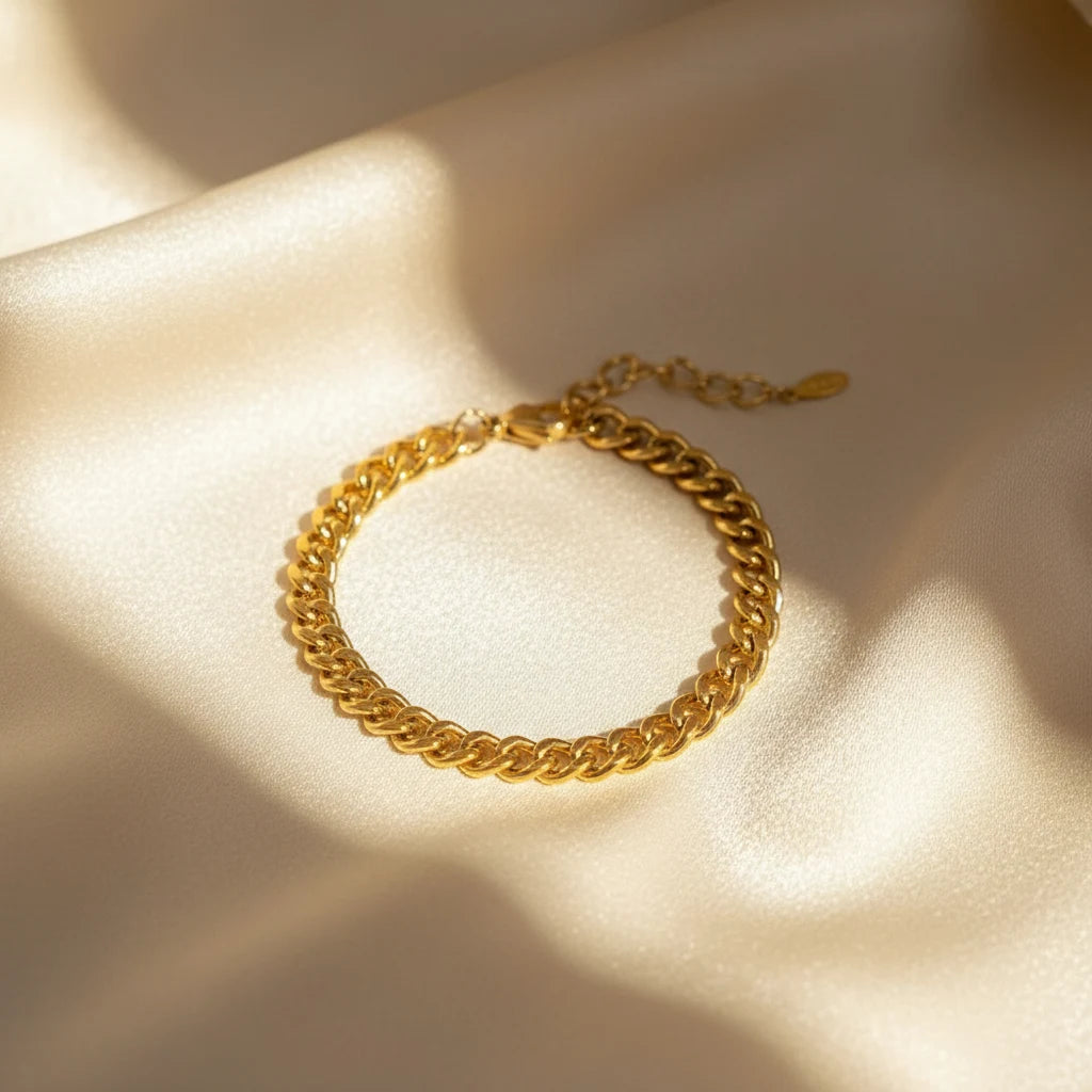 Shiny gold curb chain bracelet displayed in a circle on soft champagne satin fabric with warm sunlight and shadows | Oria Jewel