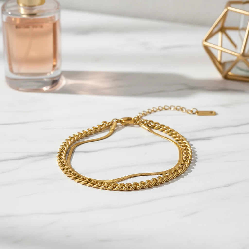 Minimalist gold double chain bracelet resting on white marble beside perfume bottle, softly lit for everyday elegance | Oria Jewel