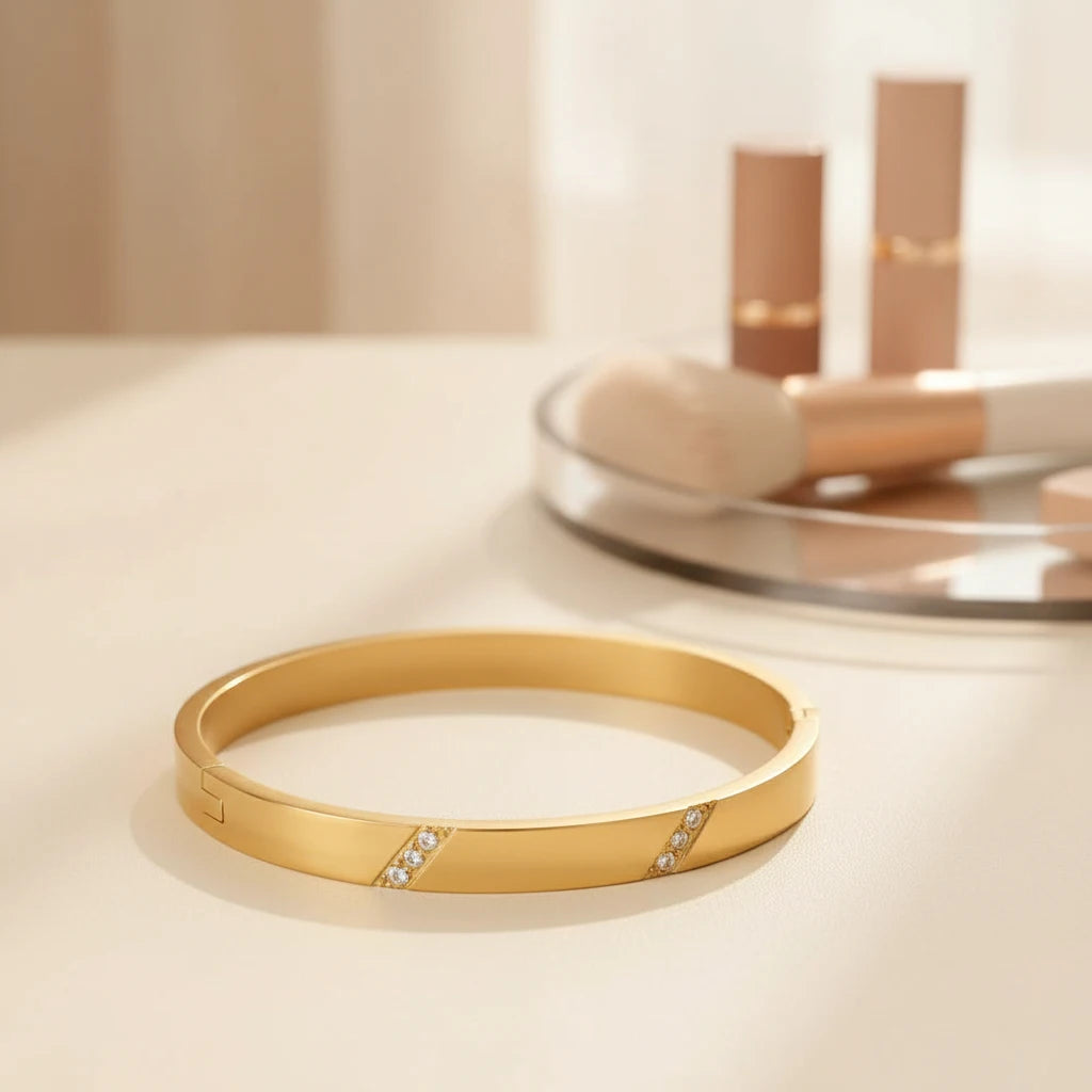 Glam vanity setup featuring a gold hinged bangle bracelet lying near nude lipsticks and a makeup brush on a mirrored tray, warm sunlight highlighting the smooth metal finish, gold hinged bangle bracelet | Oria Jewel
