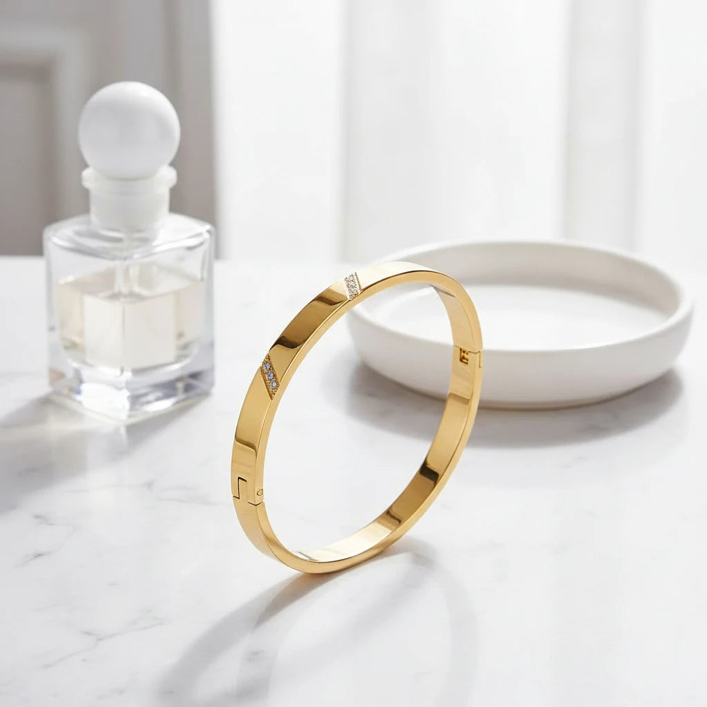 Gold hinged bangle bracelet standing upright on a white marble surface beside a clear perfume bottle and a round trinket dish, bright natural light emphasizing its sleek, modern silhouette, gold hinged bangle bracelet | Oria Jewel