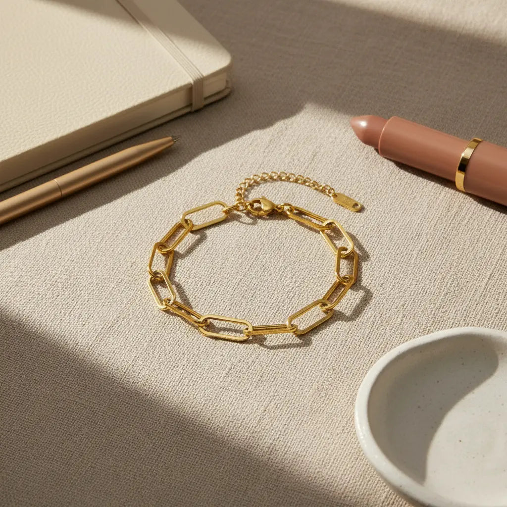 Flat lay of gold paperclip chain bracelet on textured beige fabric beside notebook, pen and lipstick for everyday styling | Oria Jewel