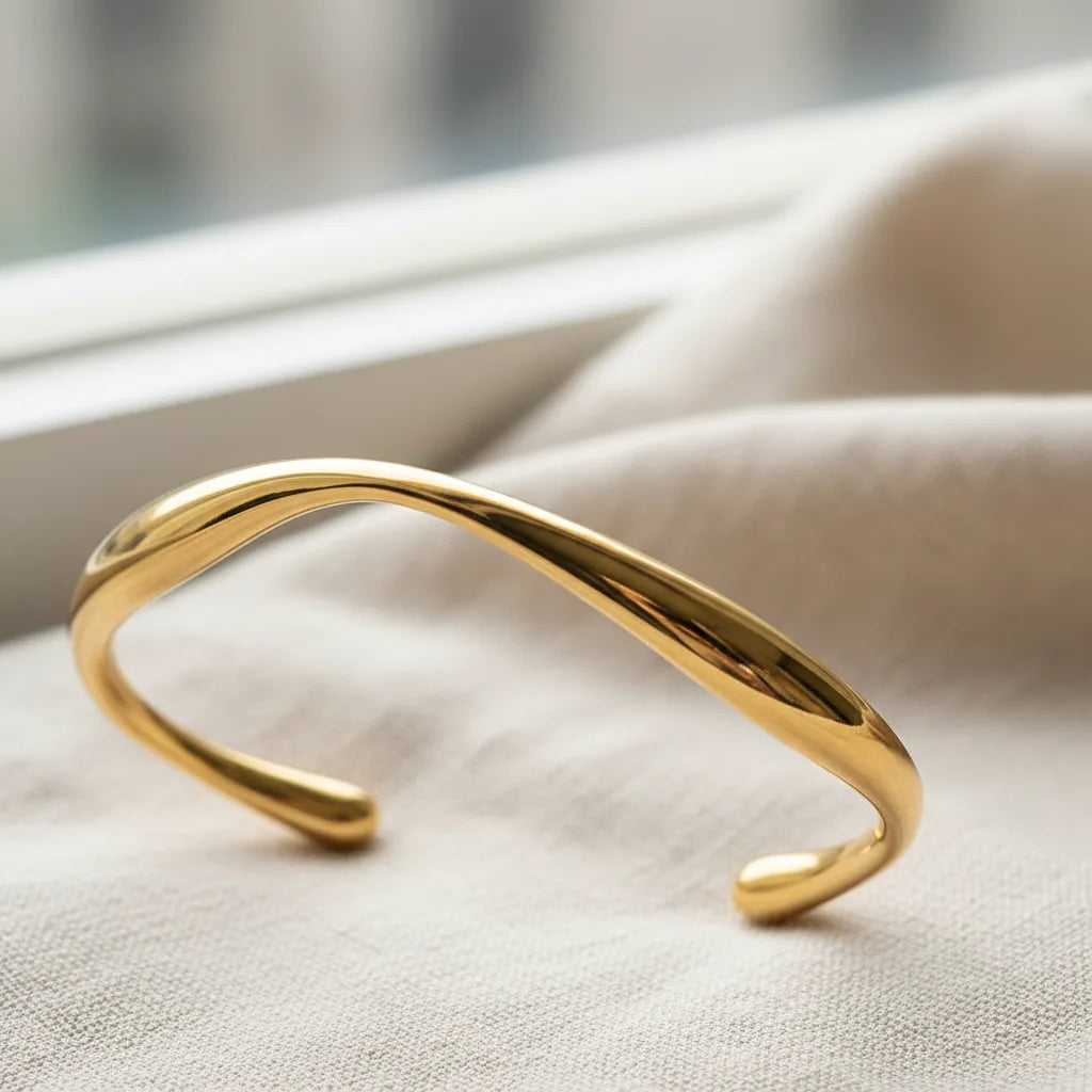 Close-up of minimalist gold cuff bracelet lying on light beige fabric near a window, polished surface reflecting soft daylight and emphasizing the fluid curve | Oria Jewel