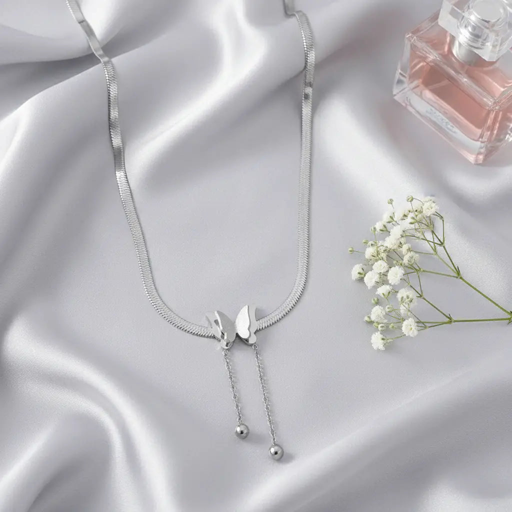 Silver Butterfly Chain Necklace arranged on soft satin fabric beside pink perfume bottle and white flowers for feminine flat lay | Oria Jewel