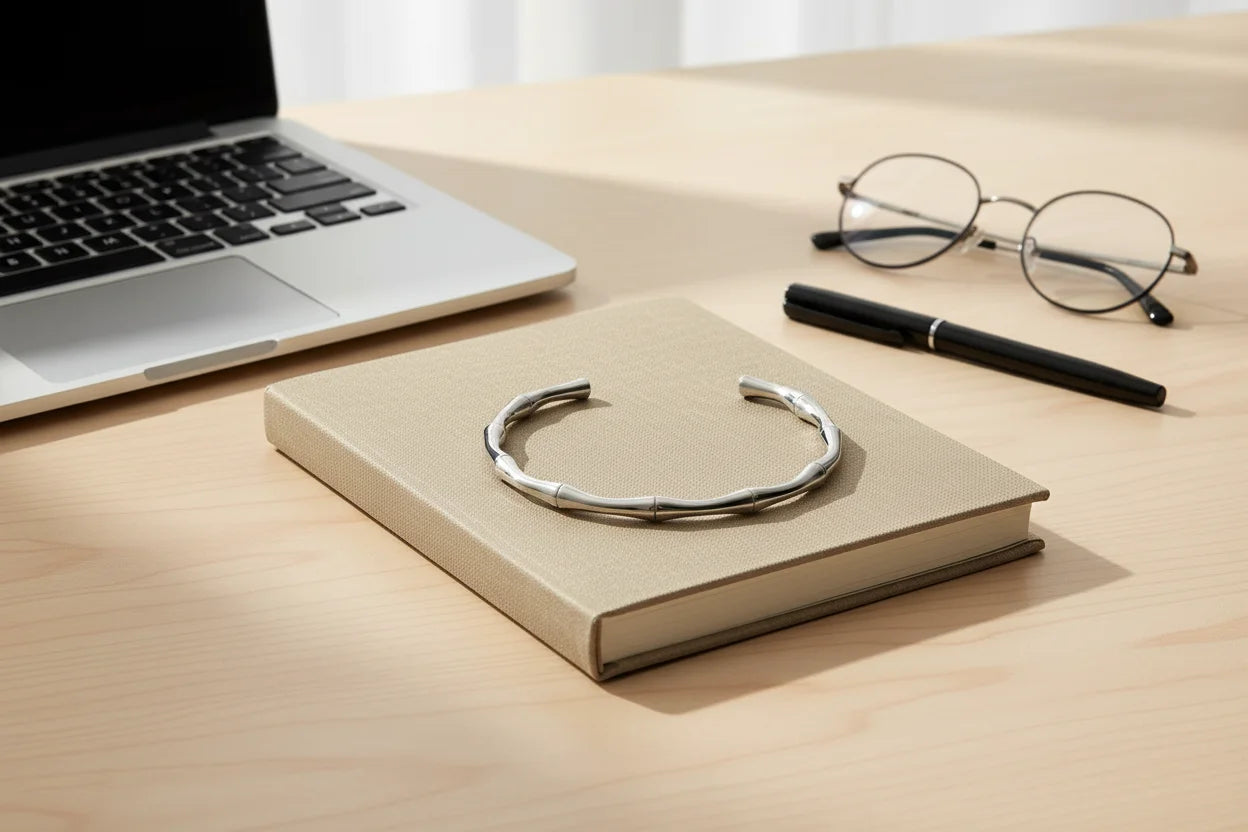 Office desk flatlay featuring a bamboo-inspired silver cuff bracelet displayed on a beige hardcover book next to a laptop, eyeglasses and black pen | Oria Jewel