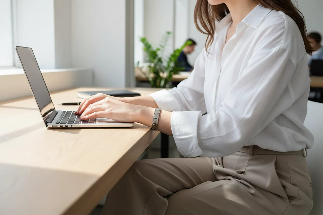 Woman typing on a laptop at a bright modern workspace, her wrist adorned with a sleek silver stainless steel stretch bracelets bracelets that adds a polished touch to her white shirt and neutral trousers | Oria Jewel